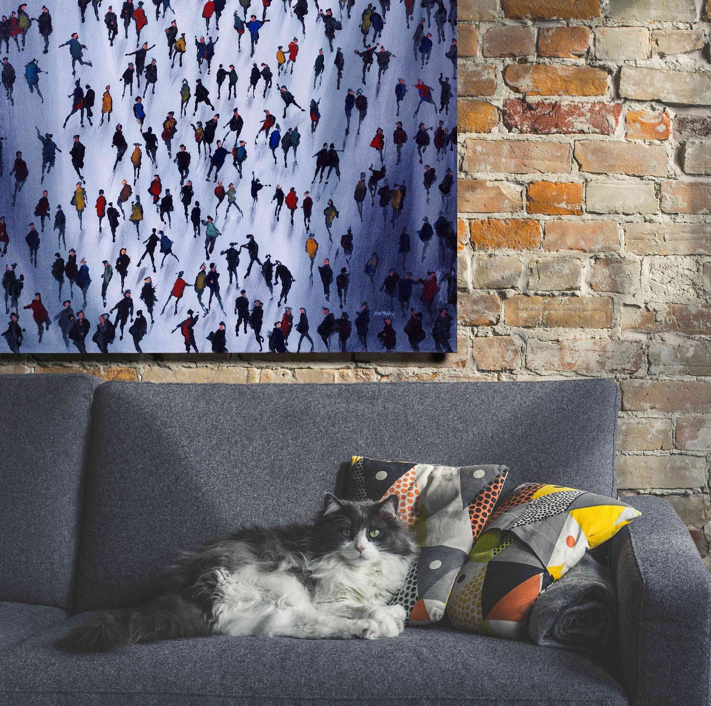 Cat lying on a grey sofa with colourful patterned cushions. A large crowd painting by Neil McBride is displayed on a bare brick wall behind the sofa.